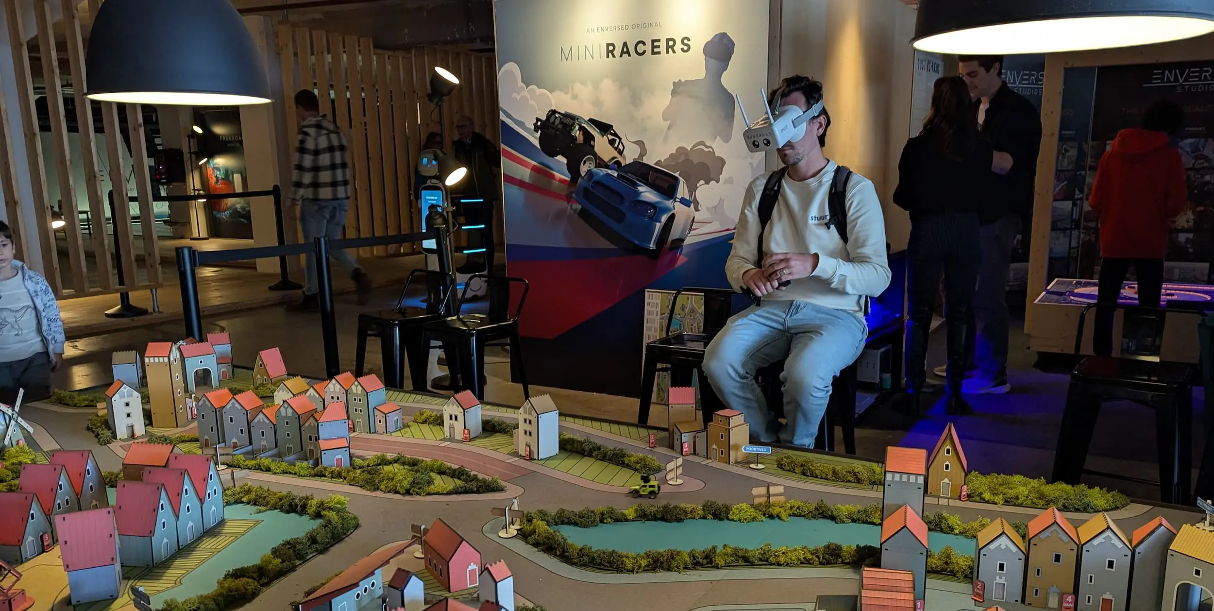 Bezoeker met een VR-headset ervaart de interactieve ‘Mini Racers’-installatie tijdens de Dutch Design Week, omringd door een kleurrijk miniatuurdorp en andere bezoekers in de expositieruimte.
